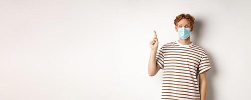 Covid, virus and social distancing concept. Surprised redhead man in face mask showing advertisement on top, pointing finger up and look amazed, white background photo