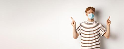 Covid, virus and social distancing concept. Handsome young man with red hair, wear face mask and pointing sideways at two promo offers, white background photo