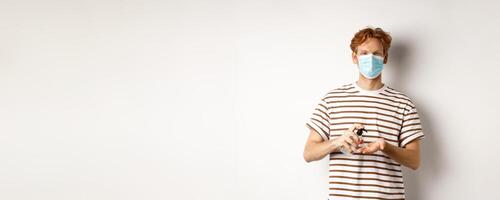 Covid, virus and social distancing concept. Teenager redhead guy in face mask clean hands with sanitizer, using antiseptic, standing over white background photo