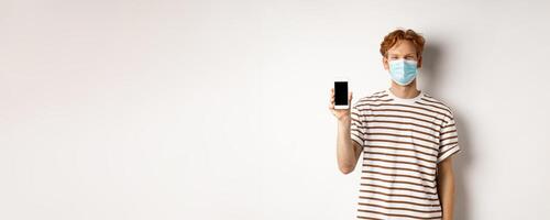 Covid-19, pandemic and social distancing concept. Handsome young man with face mask, showing black smartphone screen and smiling, standing over white background photo