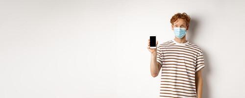 Covid-19, online doctor concept. Smiling redhead man in face mask showing smartphone screen, standing over white background photo