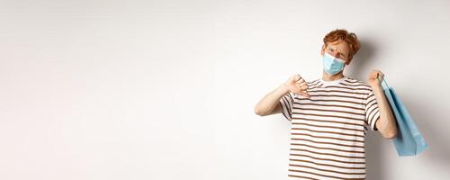 Concept of social distancing and shopping. Disappointed young man with red hair, wearing face mask, holding shopping bag and showing thumb-down from dislike, disapprove store photo