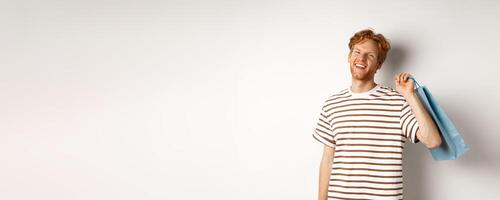Cheerful bearded man with red hair enjoying shopping, holding bag over shoulder and smiling, standing over white background photo