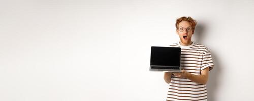 Excited redhead man drop jaw, showing laptop screen logo or website, demonstrate promo offer on display, standing against white background photo