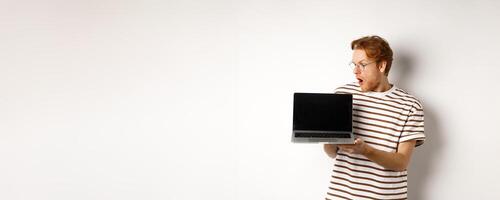 Young redhead man look impressed, showing laptop empty screen with logo banner. Guy in glasses checking out online advertisement on computer, white background photo