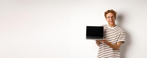 Funny redhead man in glasses showing laptop screen advertisement and smiling. Guy with red hair demonstrates promo or banner on display, white background photo