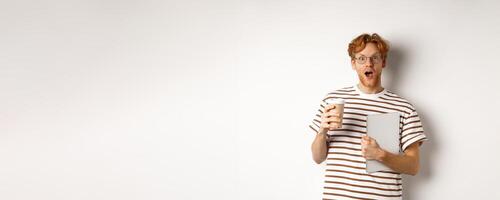 Amazed redhead man talking with coworkers on coffee break, holding cup and laptop, staring surprised at camera, white background photo