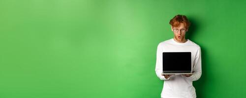 Impressed redhead nerdy guy in glasses showing blank laptop screen and looking at display amazed, standing over green background photo