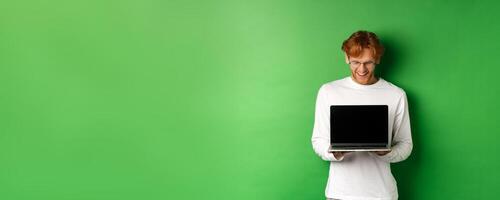 Happy young man with red hair, showing blank laptop screen and looking pleased, smiling while staring at computer, green background photo