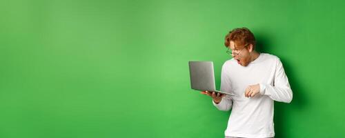 Image of young redhead man looking surprised at laptop screen, drop jaw and staring at display amazed, green background photo