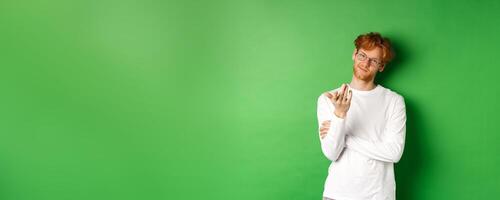 Young man with red messy hair and glasses, smiling pleased and pointing hand at camera, your turn gesture, standing over green background photo