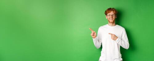 Handsome young man with red hair and beard smiling, pointing fingers left at logo, showing advertisement, standing over green background photo