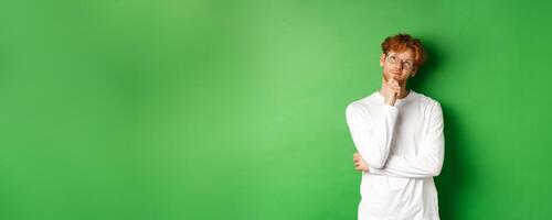Thoughtful redhead man in glasses making choice, looking up and thinking, standing over green background photo