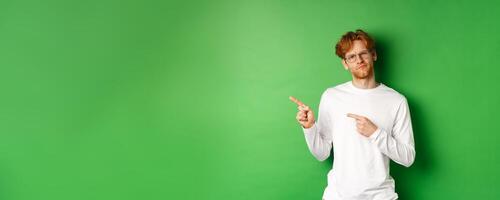 Doubtful redhead guy in glasses pointing fingers left, looking hesitant and unsure at camera, standing over green background photo