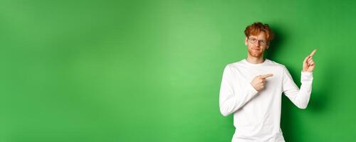 Skeptical caucasian man in glasses having doubts, pouting and frowning while pointing fingers right at something bad, standing over green background photo