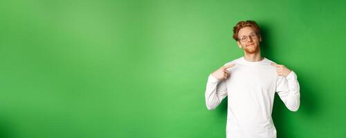 Confident and smug redhead man in glasses smiling, pointing at himself self-assured, standing over green background photo