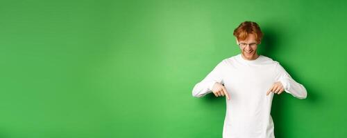 Handsome redhead man in glasses and white long-sleeve checking out advertisement, pointing fingers and looking down with pleased smile, standing over green background photo