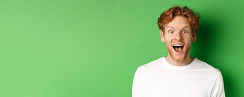Emotions and fashion concept. Close up of surprised redhead man hear amazing news, staring at camera joyful, standing over green background photo