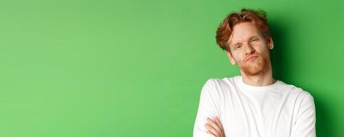 Emotions and fashion concept. Close-up of skeptical young man with red hair and beard pucker lips, looking at camera as waiting for something, standing over green background photo
