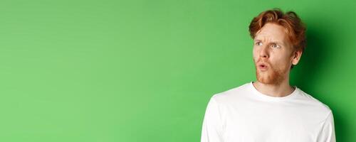 Confused redhead man staring left and looking worried, standing against green background photo