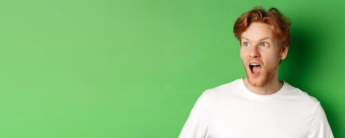 Close up of surprised and impressed redhead man checking out promotion offer, looking left with dropped jaw, standing over green background photo