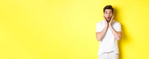Image of handsome man looking surprised, gasping and saying wow, looking at promo offer amazed, standing over yellow background photo