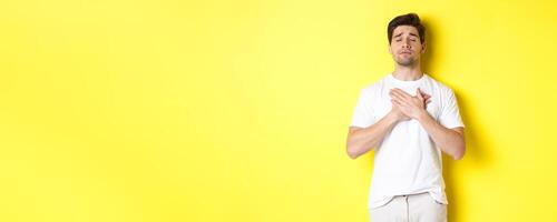Romantic and nostalgic man holding hands on heart, close eyes and remember something, standing over yellow background photo