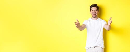 Excited handsome guy stretching hands forward, reaching for hug, receiving gift, standing over yellow background photo