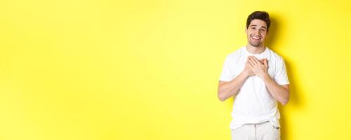 Image of grateful handsome guy in white t-shirt, holding hands on heart and smiling pleased, express gratitude, thanking for something, standing over yellow background photo