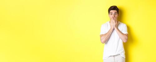 Handsome man looking shocked and speechless, holding hands on mouth, standing over yellow background photo