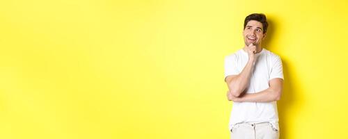 Portrait of smiling man looking thoughtful at upper left corner, choosing something, have an idea, standing against yellow background photo