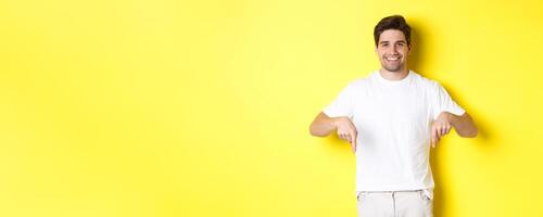 Attractive bearded man pointing fingers down, showing event banner, standing over yellow background photo