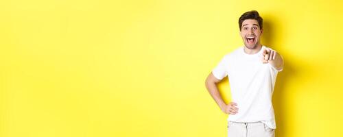 Happy guy pointing finger at camera and laughing, check out something, standing over yellow background photo