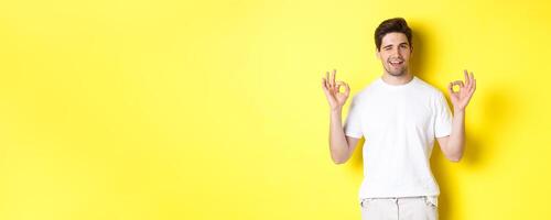 Confident handsome man winking, showing okay signs in approval, like something good, standing over yellow background photo