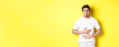Man having stomach ache, grimacing from pain and touching belly, standing over yellow background photo