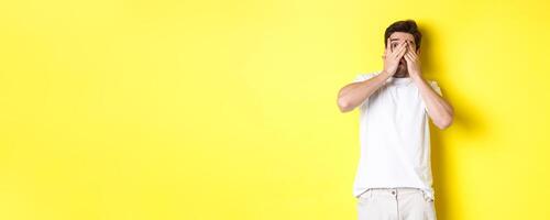 Embarrassed guy shut eyes but peeking through fingers at something awkward, standing over yellow background photo