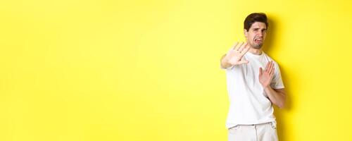 Disgusted man refusing, grimacing from dislike and aversion, begging to stop, standing in white t-shirt against yellow background photo