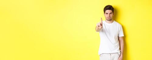 Confident man saying no, decline and prohibit something, showing one finger and frowning, standing over yellow background photo