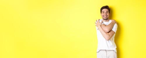 Happy kind guy hugging himself and smiling, standing pleased against yellow background photo