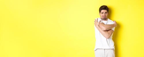 Silly and timid guy trying to comfort himself, hugging his body and frowning scared, standing over yellow background photo