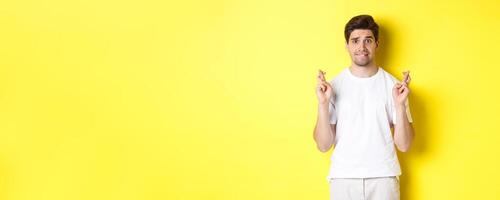 Nervous guy crossing fingers for good luck, hoping for something, standing over yellow background photo