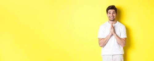 Hopeful man asking for help, begging favour, need something and smiling, standing over yellow background photo