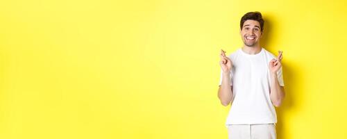 Hopeful smiling man holding fingers crossed, making wish or praying, standing over yellow background photo