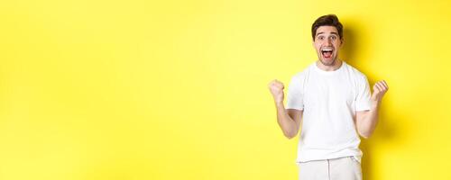 Excited and lucky man winning, clench fists and looking happy, triumphing and celebrating, standing over yellow background photo