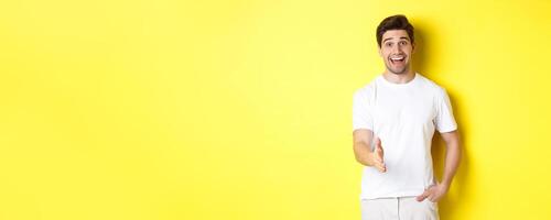 Friendly man greeting you with handshake, smiling amused, saying hello, standing over yellow background photo