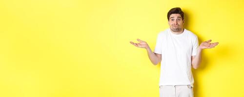 Clueless man shrugging, spread hands sideways confused, dont know anything, standing over yellow background photo