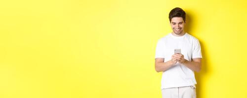 Young man reading text message on smartphone, looking at mobile phone screen and smiling, standing in white t-shirt against yellow background photo