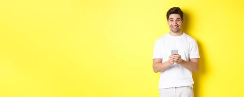 Man smiling and looking happy after reading promo offer on smartphone, standing against yellow background in white t-shirt photo