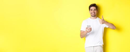 Happy satisfied man holding smartphone, showing thumb up in approval, recommend something online, standing over yellow background photo