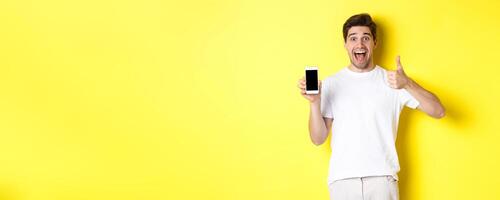 Happy young man showing thumb up and mobile phone screen, recommending application or internet website, standing over yellow background photo